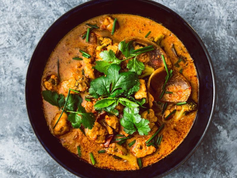 Draufsicht: Auf einem grauen Untergrund steht eine schwarze Schüssel Tofu-Curry mit Brokkoli und Koriander.