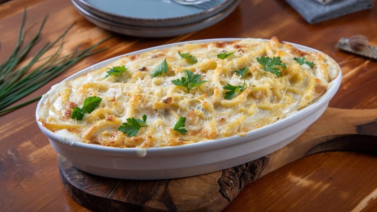 Eine weiße Auflaufform mit den überbackenen Spätzle auf einem Holzbrett und einem Holztisch mit zwei Tellern, Gabeln und einem Bund Schnittlauch.