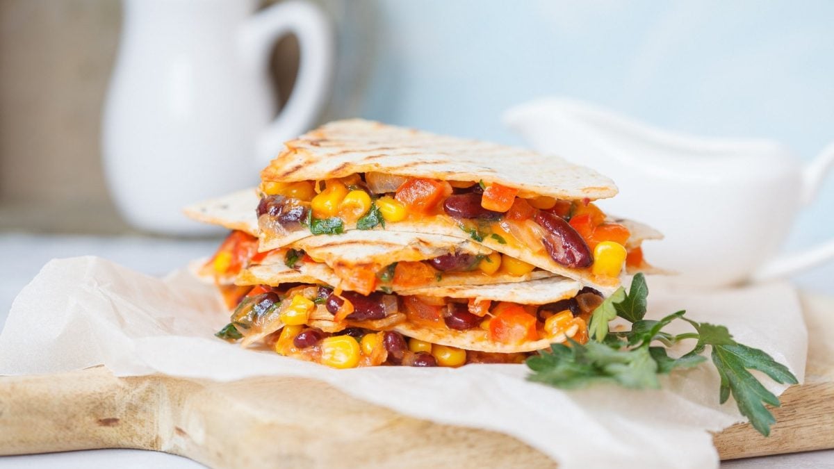 Ein Stapel vegane Quesadillas mit Mais, Paprika, Bohnen und mehr, auf Butterbrotpapier auf einem Brett.