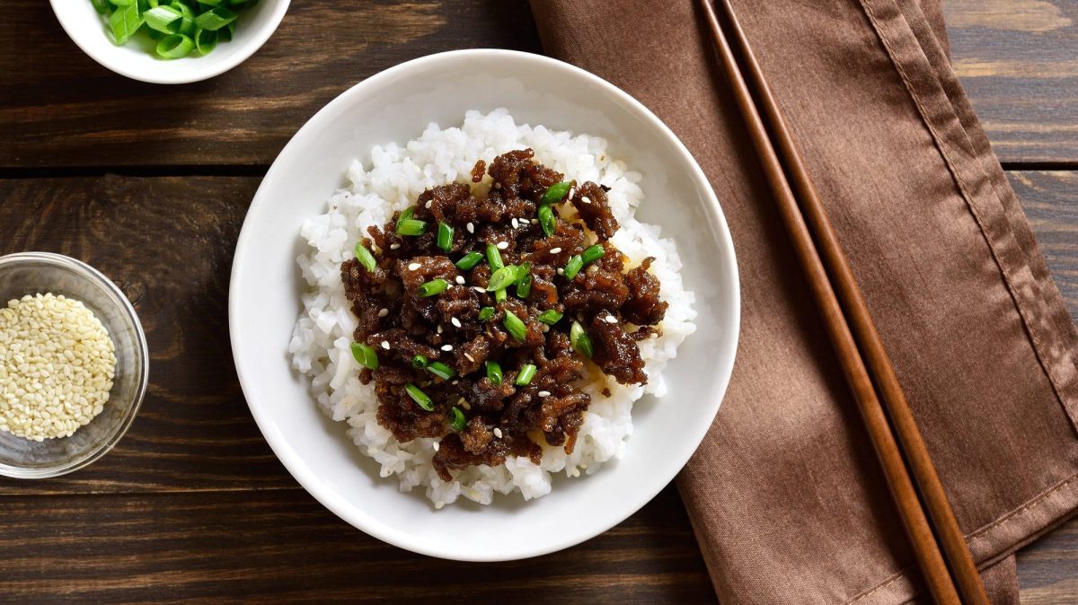 Ein Teller Reis und veganes Bulgogi mit Frühlingszwiebeln und Sesam bestreut in der Draufsicht, daneben Essstäbchen und Schalen mit Sesam und Frühlingszwiebeln.