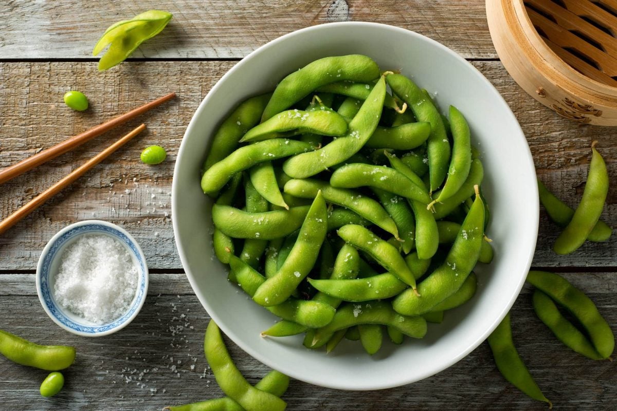 Draufsicht: Frisch gedämpfte Edamame mit Meersalz auf einer rustikalen Tischplatte bestreut.