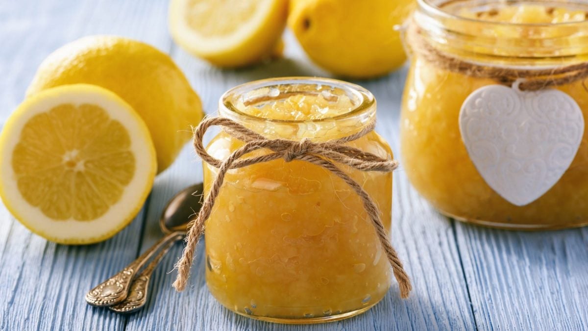 Zwei Gläser Zitronenmarmelade neben Zitronenhälften auf blauem Untergrund.