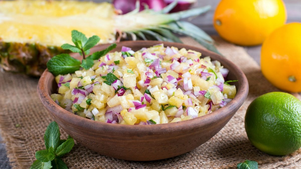 Eine braune Schale mit der Ananas-Salsa auf eionem Leinentuch mit Früchten im Hintergrund.