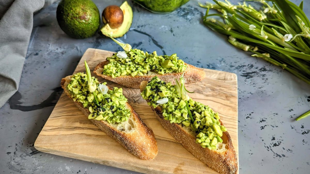 Bagutte mit dem Bärlauch-Avocado-Aufstrich auf einem Holzbrett mit dunkelgrauem Untergrund. Im Hintergrund Avocado und Bärlauch.