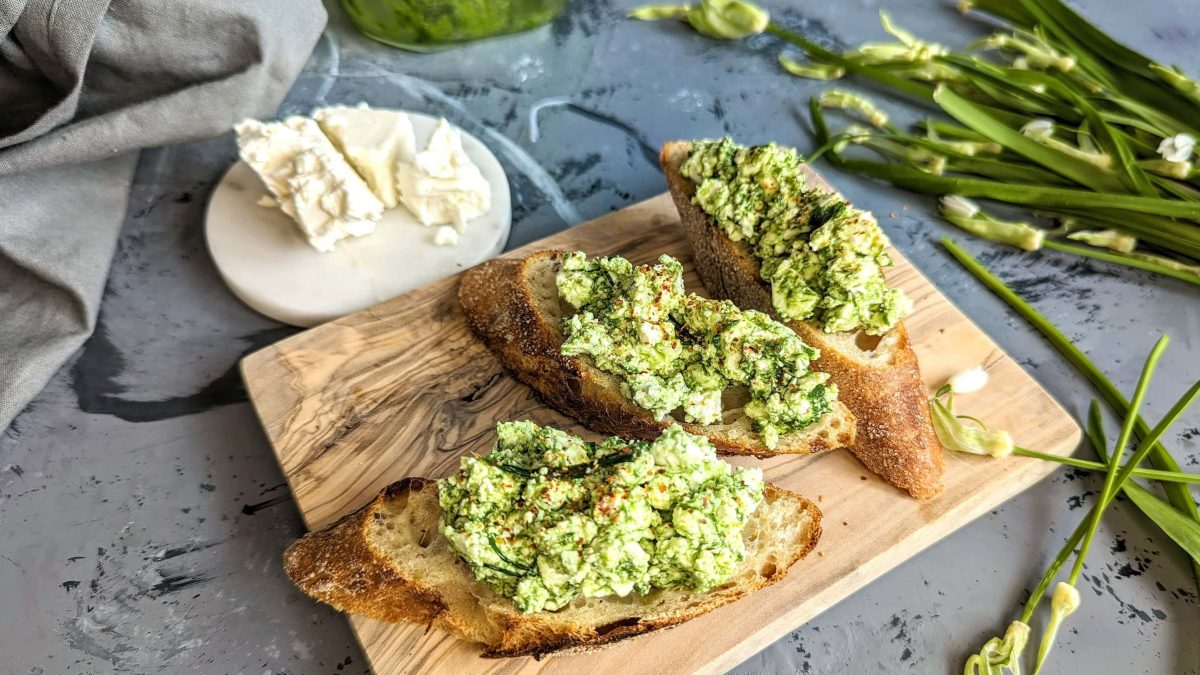 Ein kleines Holzbrett mit Baguettescheiben und der Bärlauch-Feta-Creme auf einem grauen Tisch mit Feta und Bärlauch im Hintergrund.