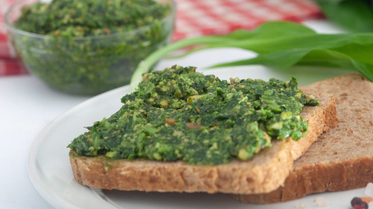 Frontalsicht: Ein Toast mit Bärlauch-Sardellen-Aufstrich liegt auf einem Holzbrettchen. Dahinter liegen Bärlauchblätter, sowie ein rot-weiß-kariertes Tuch.