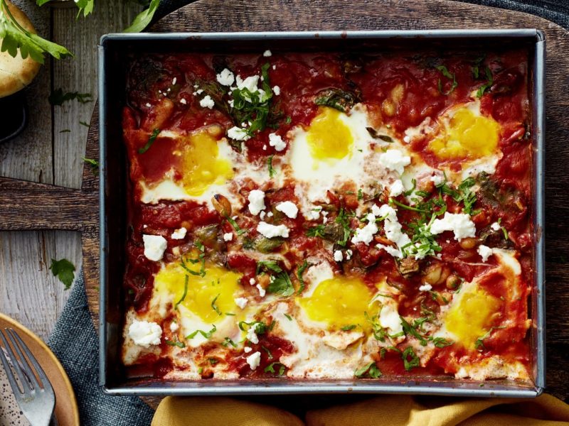 Eine Backform mit Ofen-Shakshuka mit Spinat auf einem gedeckten Tisch.