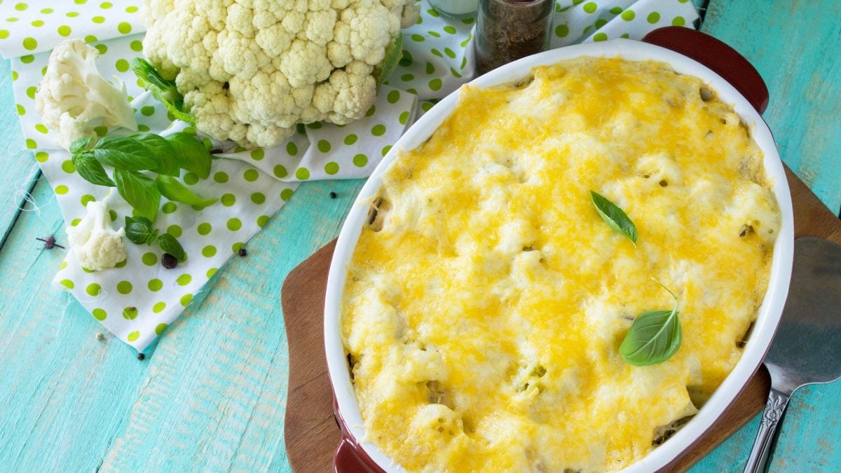Draufsicht: Auf einem blauen Holztisch liegt ein Brettchen. Auf dem liegt eine ovale Auflaufform mit einem Blumenkohl-Lachs-Gratin mit Appenzeller. Neben der Form ist frischer Blumenkohl sowie eine Gabel.