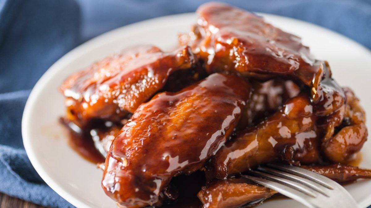 Ein weißer Teller mit den Cola-Chicken-Wings auf einem dunklen Holztisch mit einem blauen Tuch.