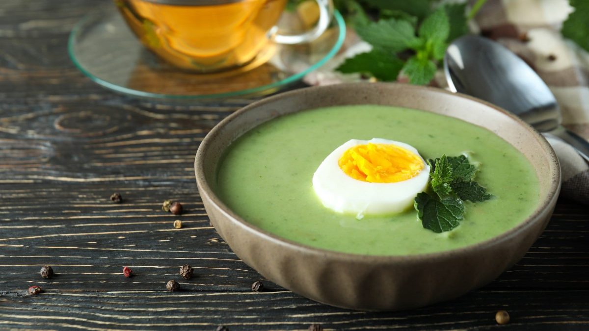 Draufsicht, schräg: Auf einem dunklen Untergund stehe eine Schüssel Brennnessel-Suppe mit Ei. Dahinter ist eine Tasse aus Glas mit einer goldigen Flüssigkeit drin.