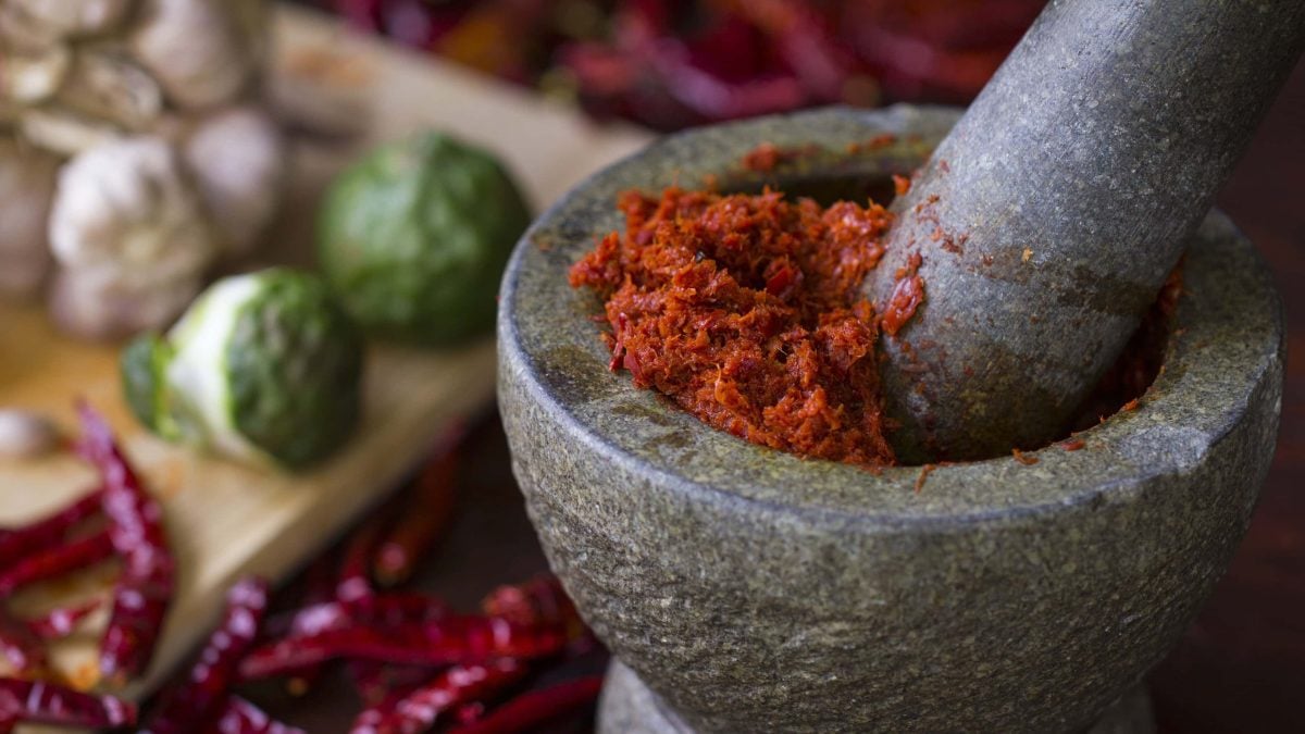 Ein Steinmörser mit Chilischoten, Limetten und Knoblauch unscharf im Hintergrund. Im Steinmörser eine rote Currypaste.