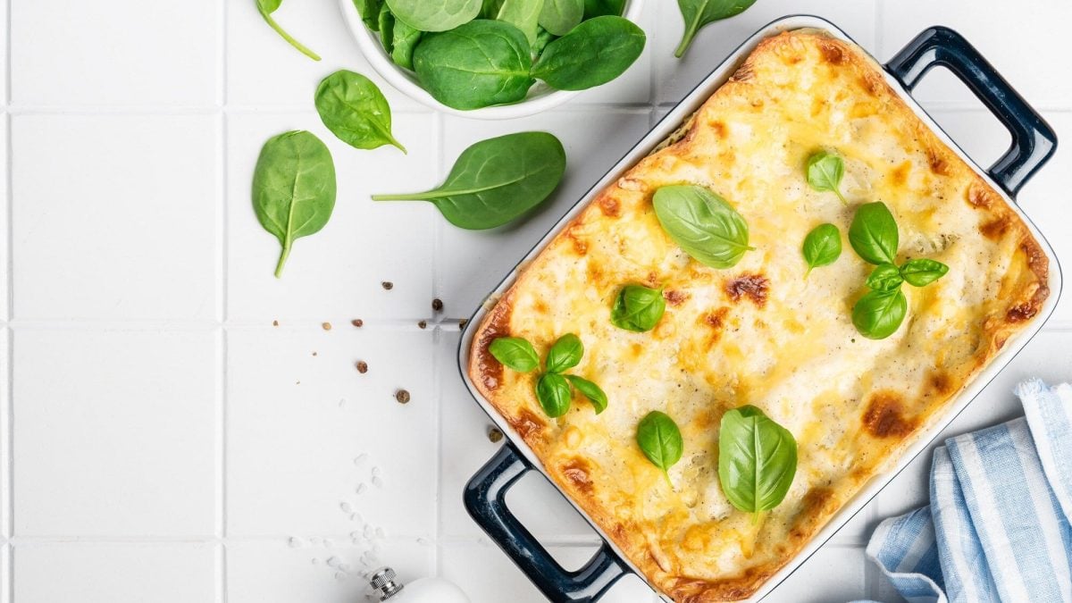 Draufsicht: Auf einem weißen Untergrund steht eine Spinat-Basilikum-Lasagne. Drumherum und obenauf liegen Spinat- und Basilikum-Blätter.