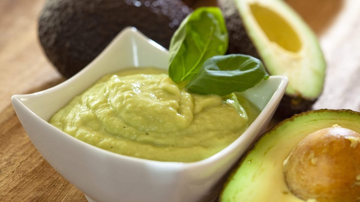 Feta-Joghurt-Creme mit Basilikum und Avocado in weißer Schale auf Holz. Im Bild Avocados und Basilikum. Draufsicht.