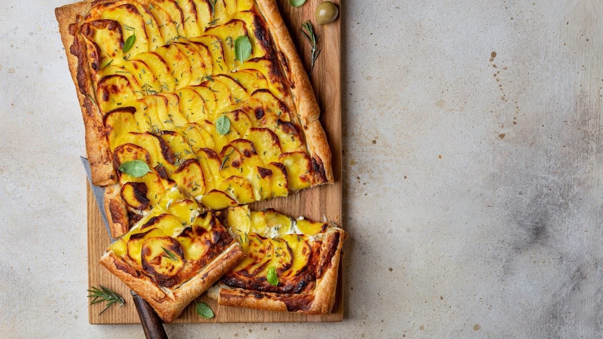 Ein Holzbrett mit dem Flammkuchen, der mit Kartoffeln und Rosmarin belegt ist auf hellem Steinuntergrund.