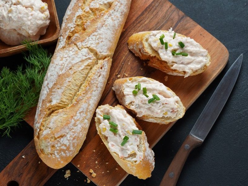 Draufsicht: Dunkler Untergrund; ein angeschnittenes Baguette sowie Baguette-Scheiben auf einem Holzbrettchen. Auf den Baguette-Scheiben ist ein Forellen-Rilette.