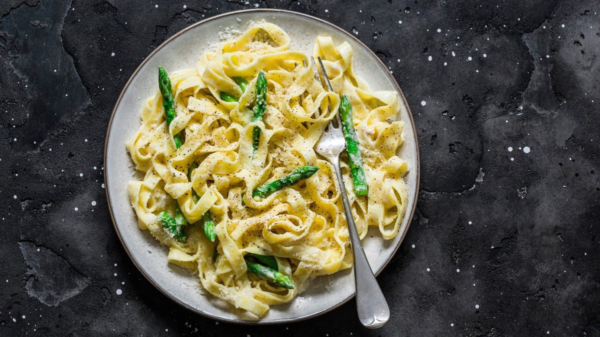 Frischkäse-Pasta mit grünem Spargel auf einem Teller, darauf eine Gabel, auf dunklem Untergrund, Draufsicht.