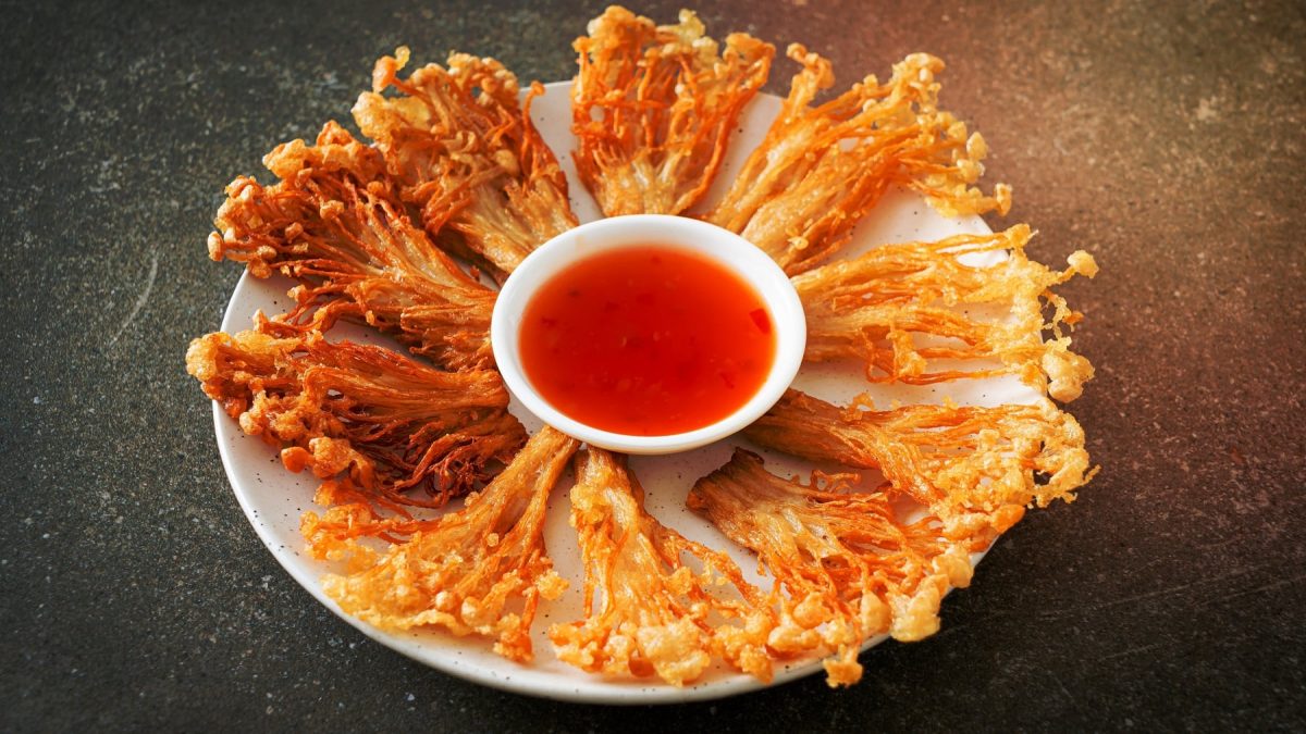 Ein weißer Teller mit den frittierten Enoki und einer kleinen Schale mit Sweet-Chili-Sauce auf einem dunklen Tisch.