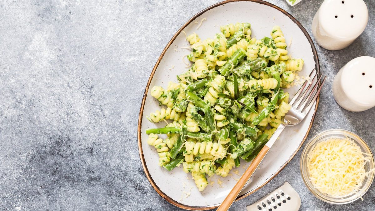 Ein ovaler, weißer Teller mit den Fusilli, grünen Bohnen und Käsesauce. Daneben Rucola, Käse, Salz und Pfeffer.