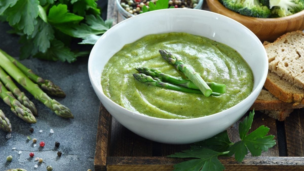 Eine Schüssel grüne Spargelcremesuppe mit Spargelstangen garniert, drumherum liegen Zutaten.