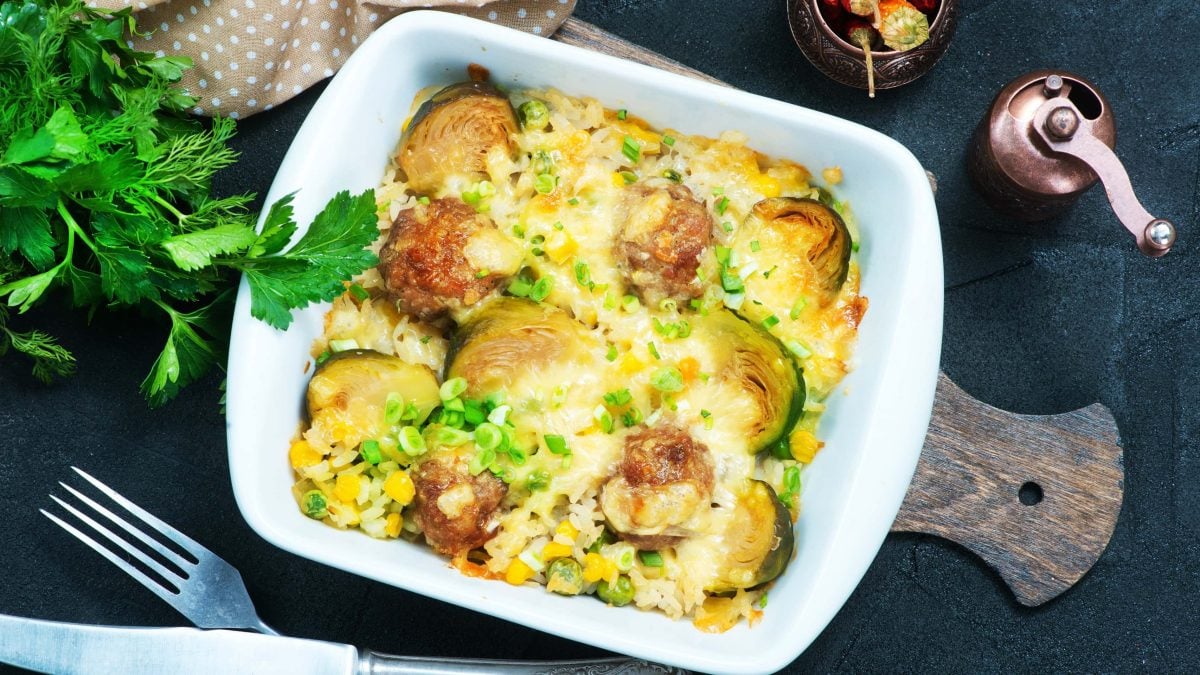 Draufsicht: Eine Hackbällchen-Zucchini-Gratin liegt auf einem dunklen Untergrund. Drum herum liegen Kräuter, Gemüse und Gewürze.