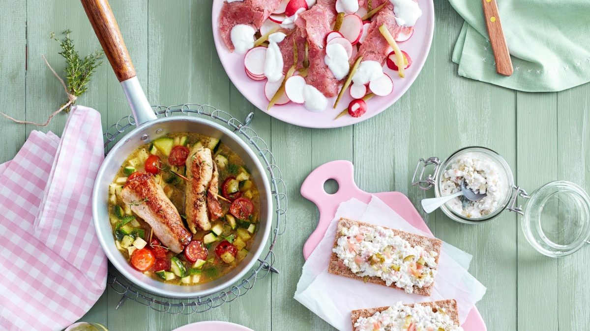 Kalbsröllchen mit Zucchini in einer Pfanne, daneben andere Gerichte, alles in der Draufsicht vor grünem Holzhintergrund.