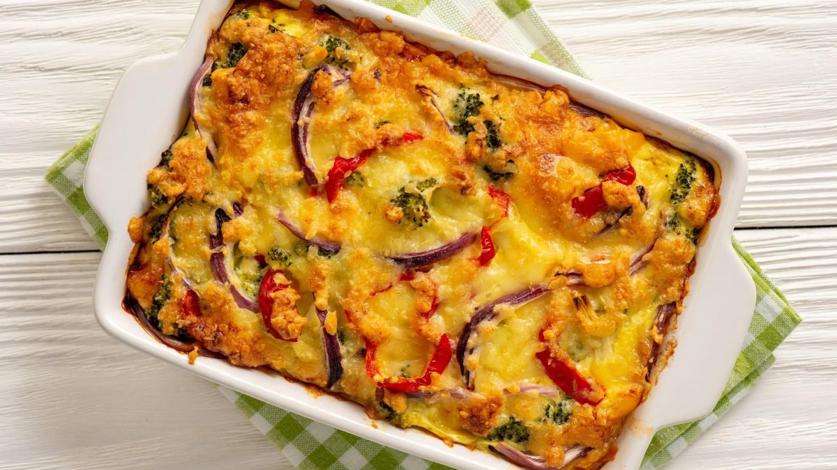 Kartoffelauflauf mit Brokkoli und Paprika in weißer Auflaufform auf weißem Holz.