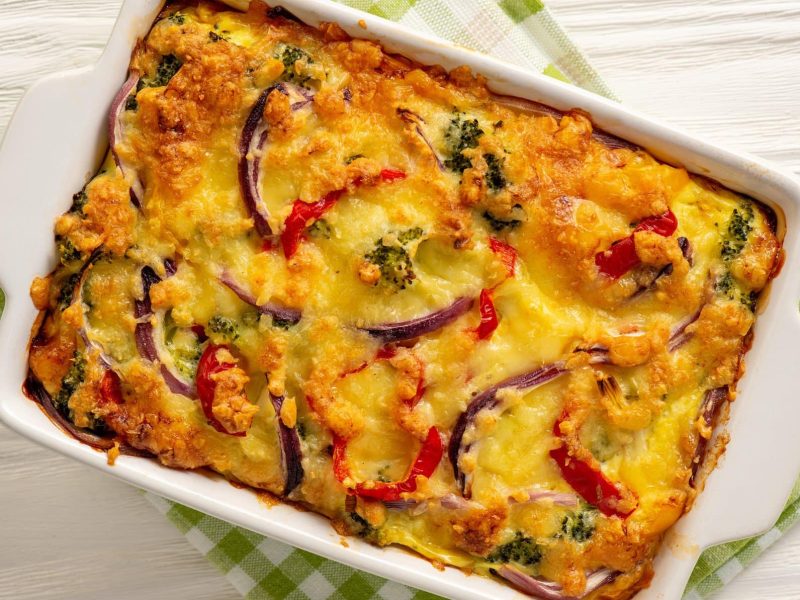 Kartoffelauflauf mit Brokkoli und Paprika in weißer Auflaufform auf weißem Holz.