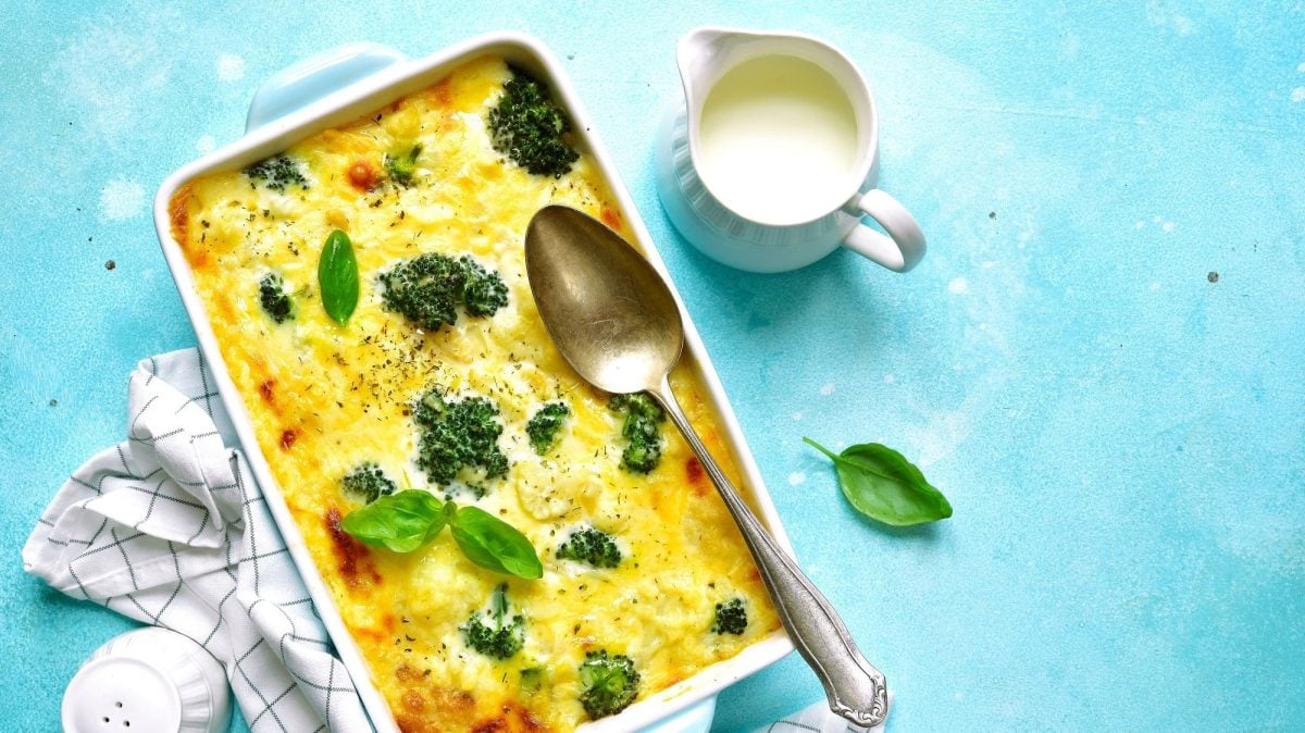 Eine Ofenform mit Kohlrabi-Brokkoli-Auflauf mit Kartoffeln und Käse, darauf ein Löffel, daneben ein Kännchen Sauce und ein Salzstreuer. Alles Draufsicht.