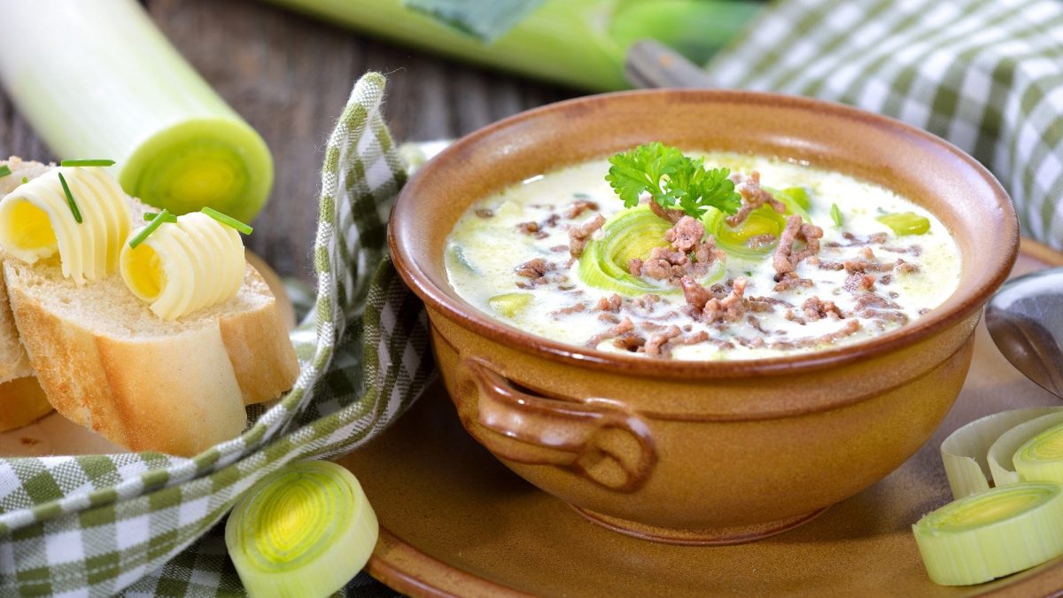 Frontalsicht: In einer runden Tonschale ist eine Hackfleisch-Suppe mit Porree. Drumherum liegen Lauch, Weißbrotscheiben und ein kariertes Küchentuch