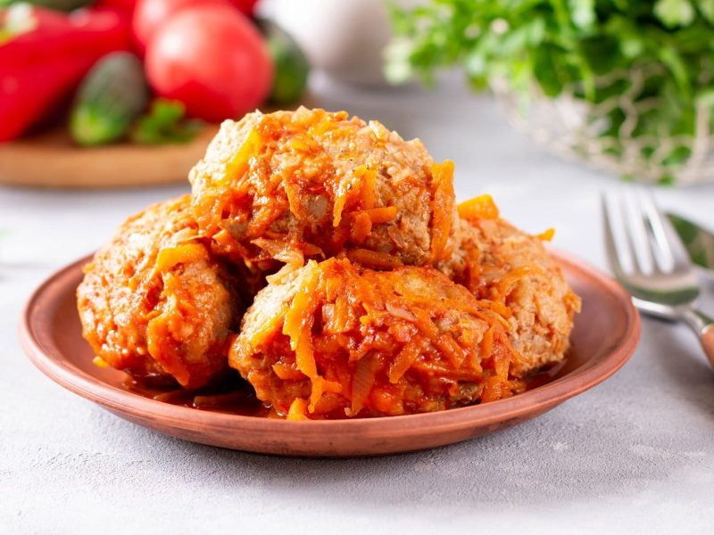 Vier ungewickelte Kohlrouladen auf einem braunen Teller und hellgrauem Untergrund. Im Hintergrund Tomaten und Paprika.