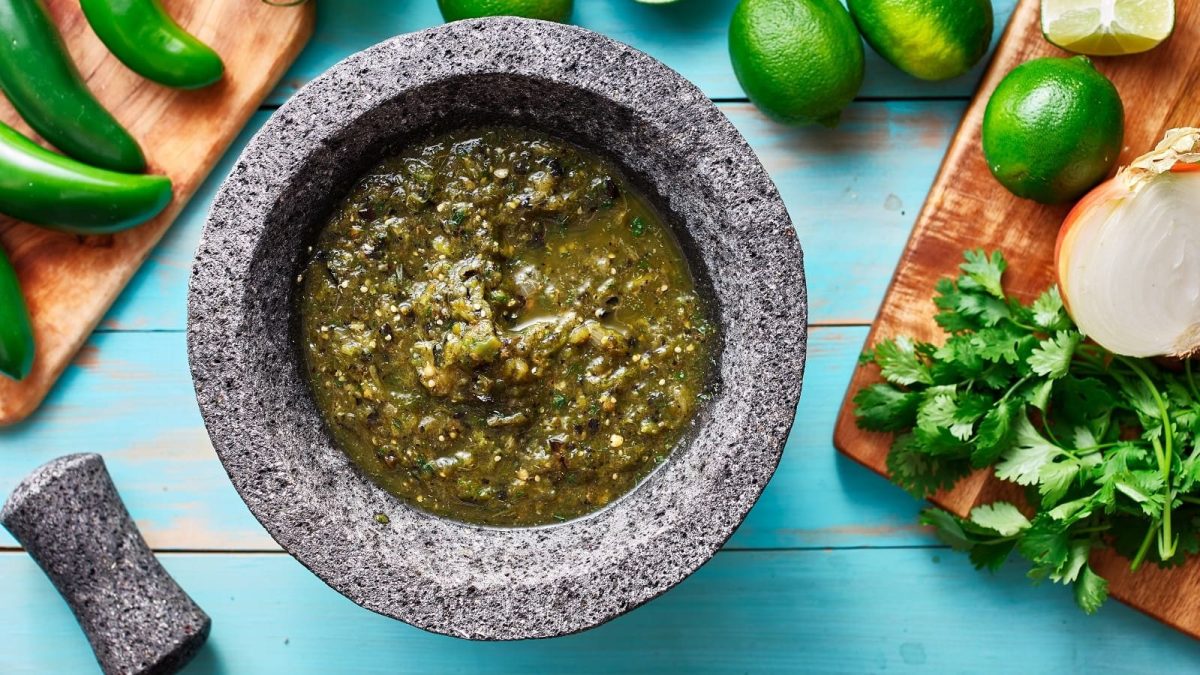Ein Steintopf mit Limetten-Koriander-Salsa auf einem blauen Tisch neben frischen Zutaten.