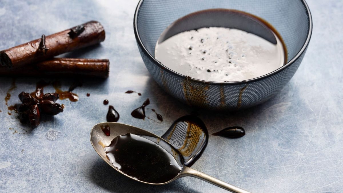 Masala Chai Sirup in blauer Schale auf grauem Untergrund. Im Bild ein Teelöffel, von dem Sirup herunter läuft. Oben Zimt und Sternanis. Draufsicht.