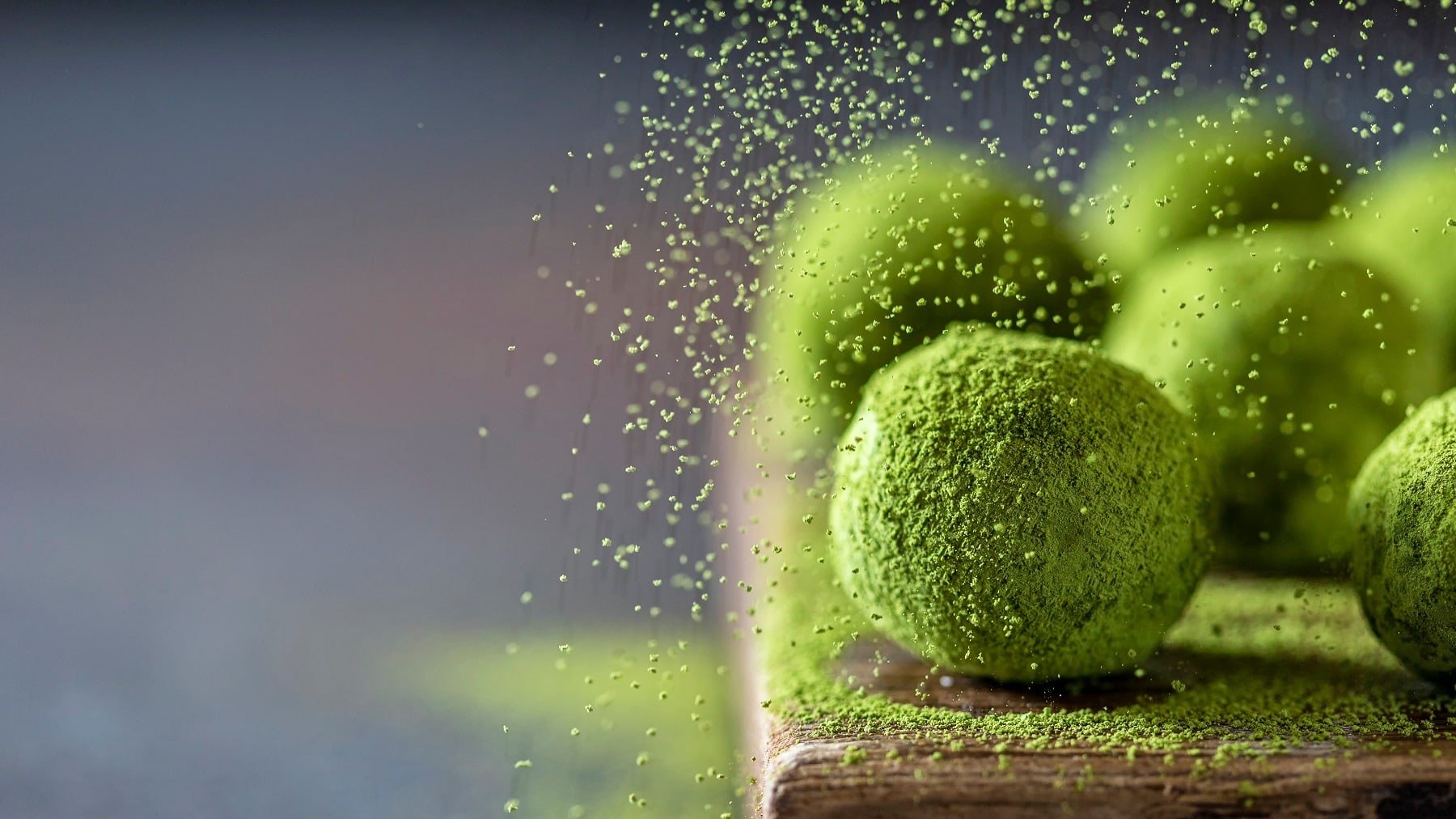 Mehrere Matcha Fudge Bällchen auf einem Holzbrett mit grauem Untergrund. Zudem wird gerade Matcha-Pulver darüber gerieselt.