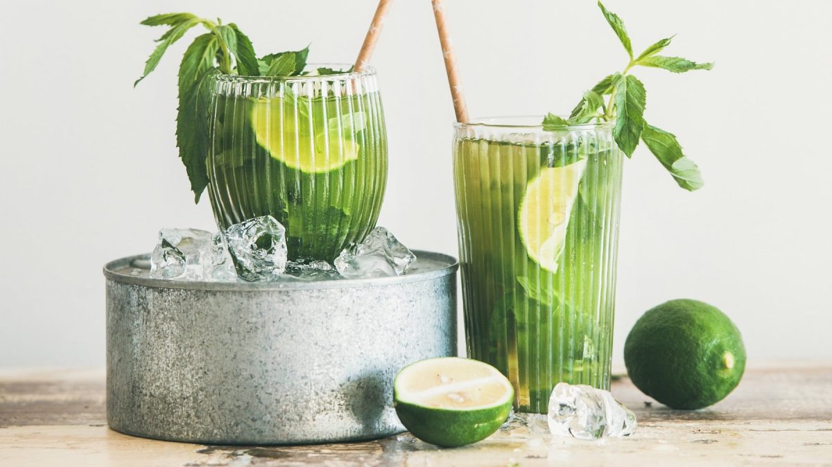 Zwei Gläser mit dem Matcha-Mocktail auf einem rustikalen Holztisch mit Eis, Limette und Minze.
