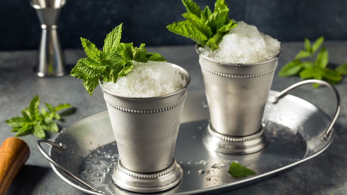 Zwei Mint Julep im Metallbecher auf Metallteller mit Minze. Im Hintergrund Barmaß, Stößel und Minze. Frontalaufnahme.