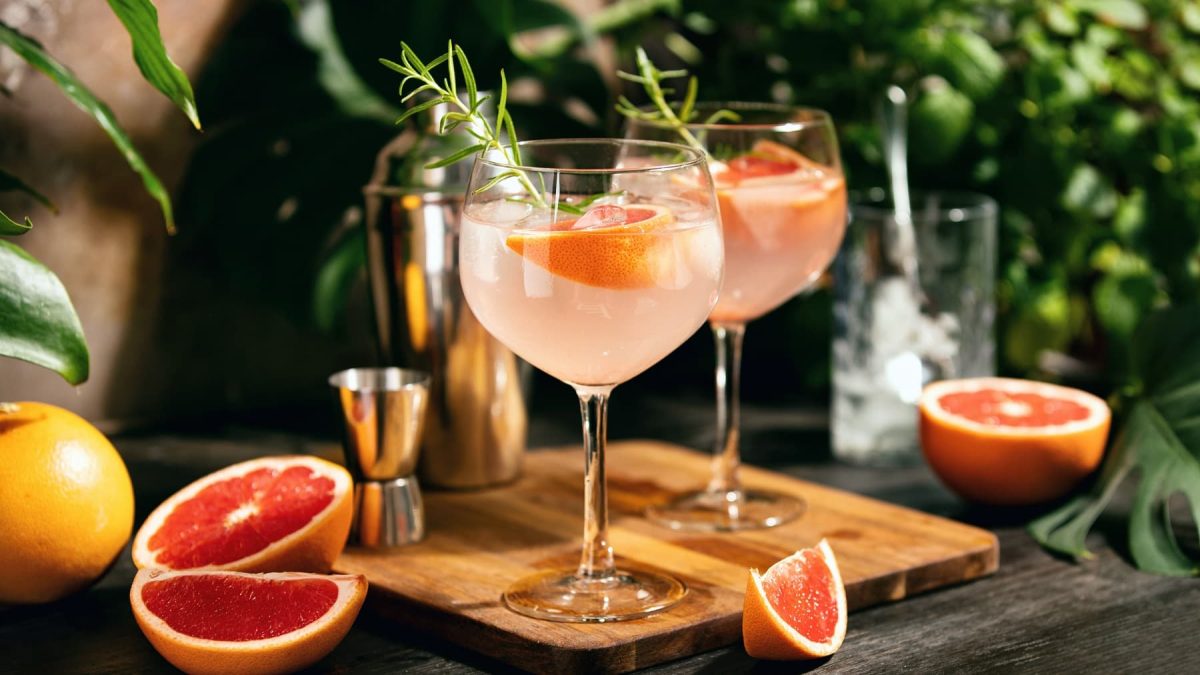 Zwei Gläser mit dem Pink Spritz auf einem Holzbrett. Im Hintergrund unscharf Pflanzen zu erkennen.