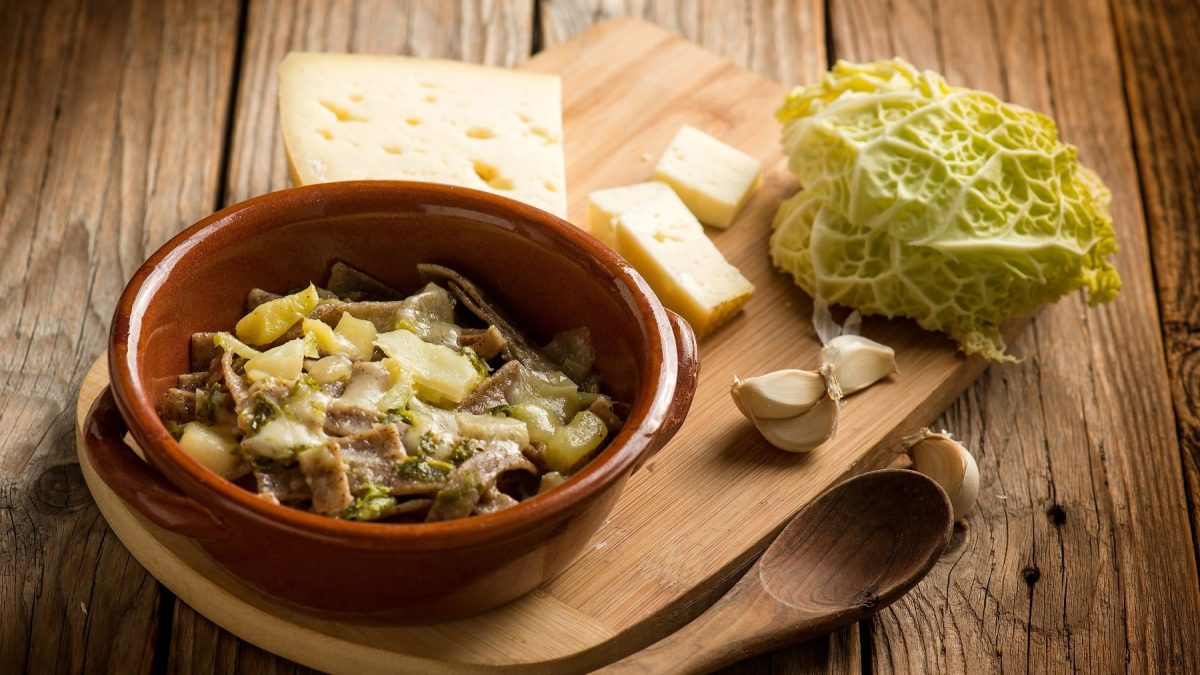 Eine braune Schale mit den Pizzoccheri-Nudeln auf einem Holzbrett mit Käse, Knoblauchzehen und etwas Wirsing.