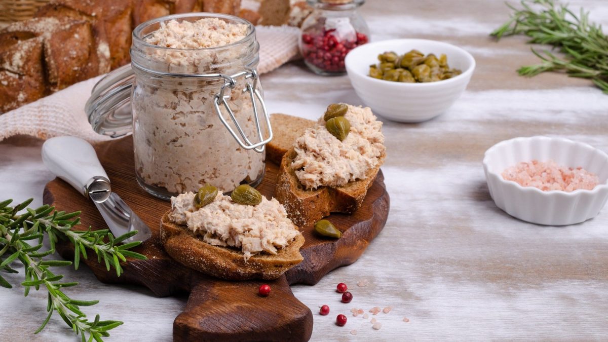 Frontal: Ein Einmachglas mit selbst gemachtem Lachs-Rillette steht auf einem dunklem Holzbrett. das liegt auf einem hellem Untergrund. Daneben lieben zwei kleine Weißbrotscheiben mit dem Lachs-Rille, Rosmarinzweige sowie eine kleine Schüssel mit Kapern.