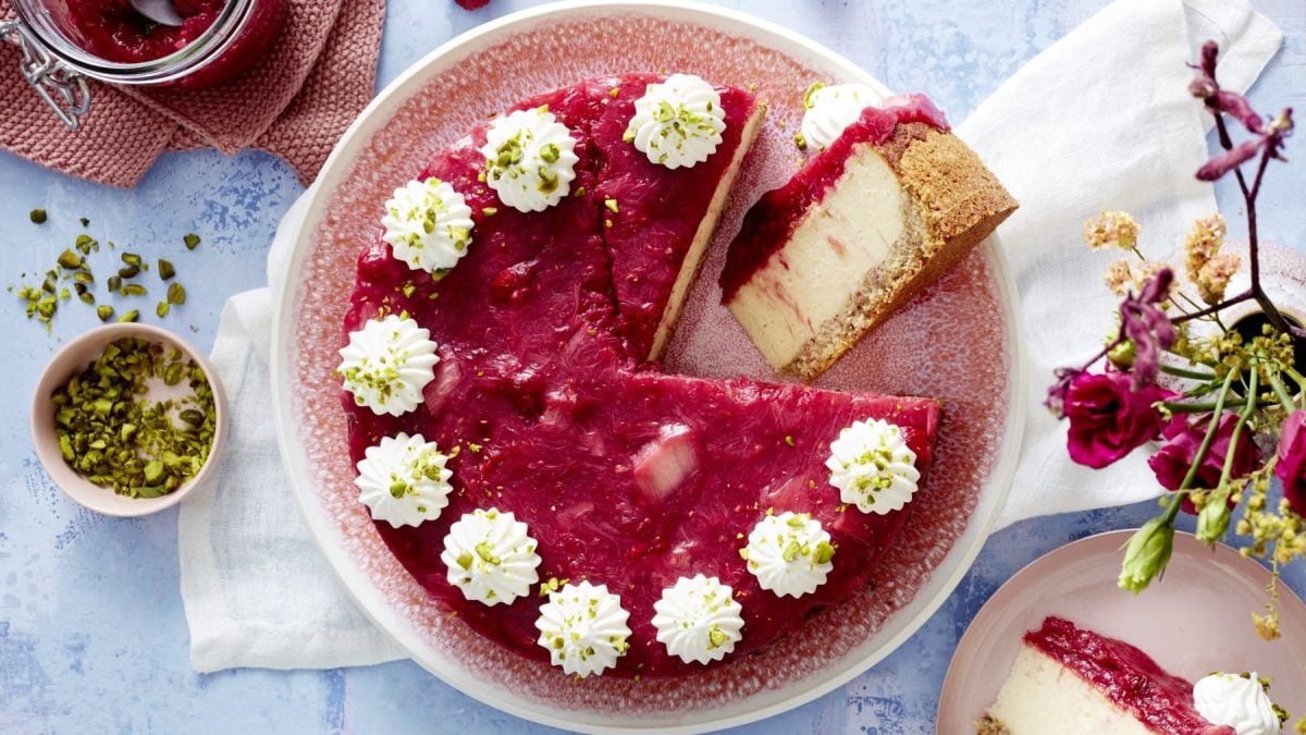 Draufsicht: Angeschnittener, runder Rhabarber-Grieß-Cheesecake mit Sahnetuffs. Daneben eine Schale mit zerbröselten Pistazien und Deko.