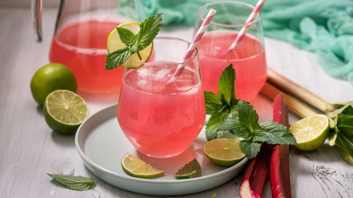 Zwei Gläser Rhabarber-Holunder-Spritz mit Strohhalmen, Minze und Limetten, drumherum die Zutaten.