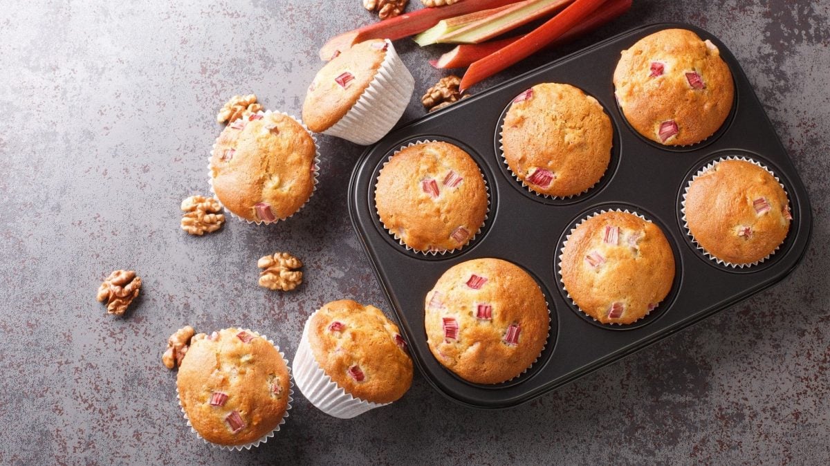 Ein Blech mit Rhabarber-Walnuss-Muffins in der Draufsicht, daneben liegen Walnusskerne, Rhabarberstangen und weitere Muffins.