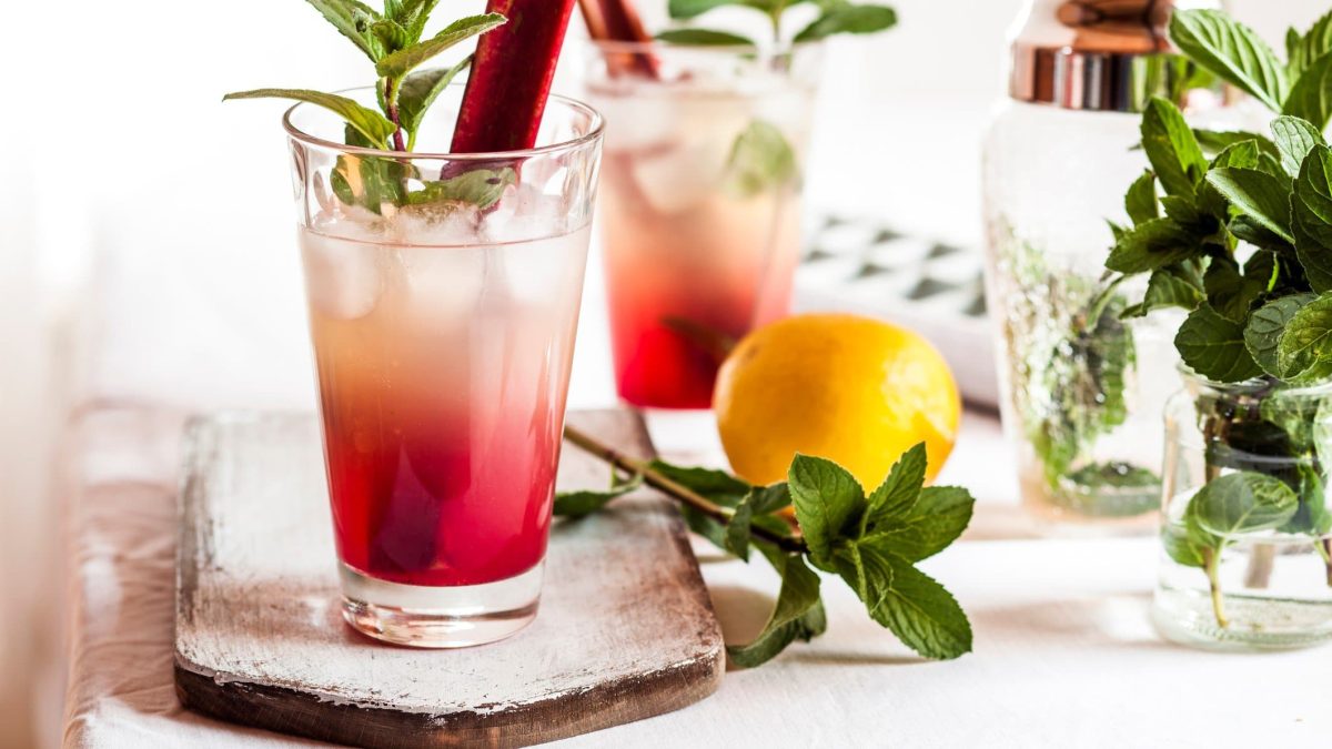 Rhabarber-Collins mit selbstgemachte Rhabarber-Limo mit Minze und Rhabarber-Stange im Glas. Im Bild ein weiterer Drink, Zutaten und Cocktail-Zubehör. Frontalansicht.