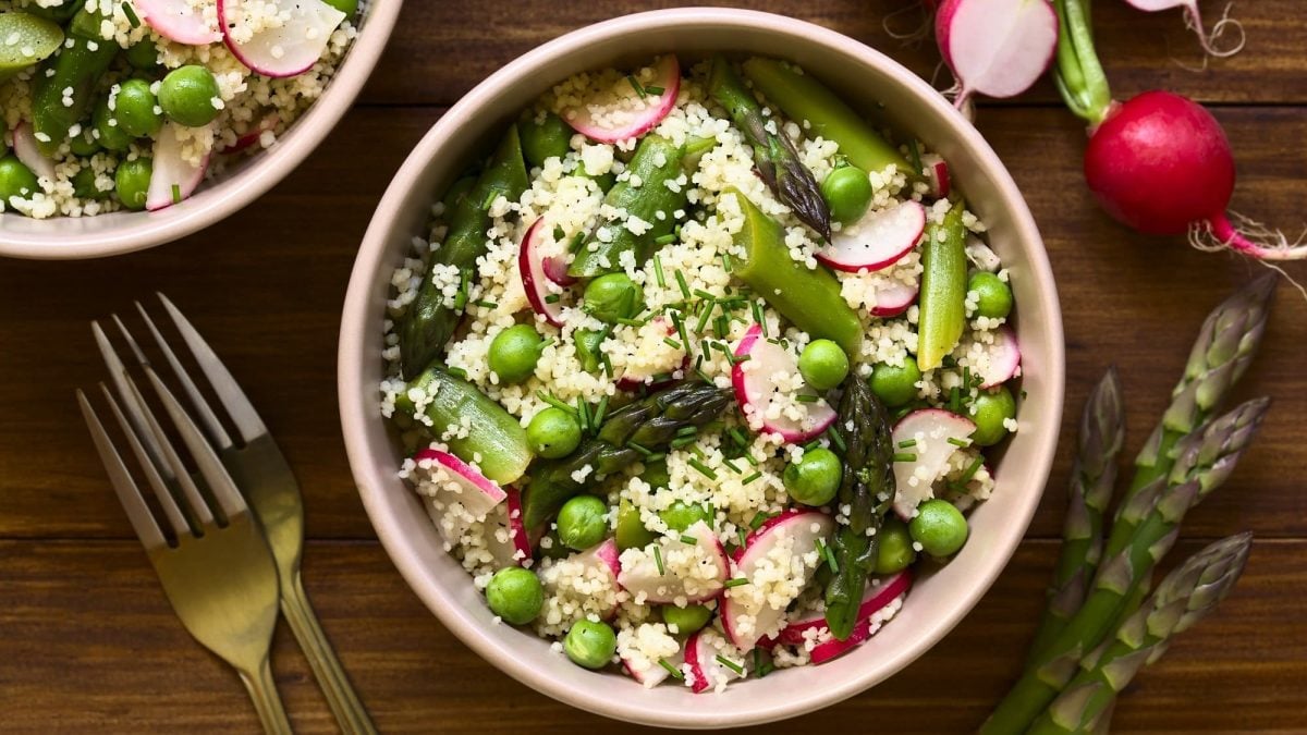 Zwei Schüsseln Spargel-Couscous-Salat mit Erbsen und Radieschen in der Draufsicht, daneben zwei Gabeln, grüne Spargelstangen und Radieschen.