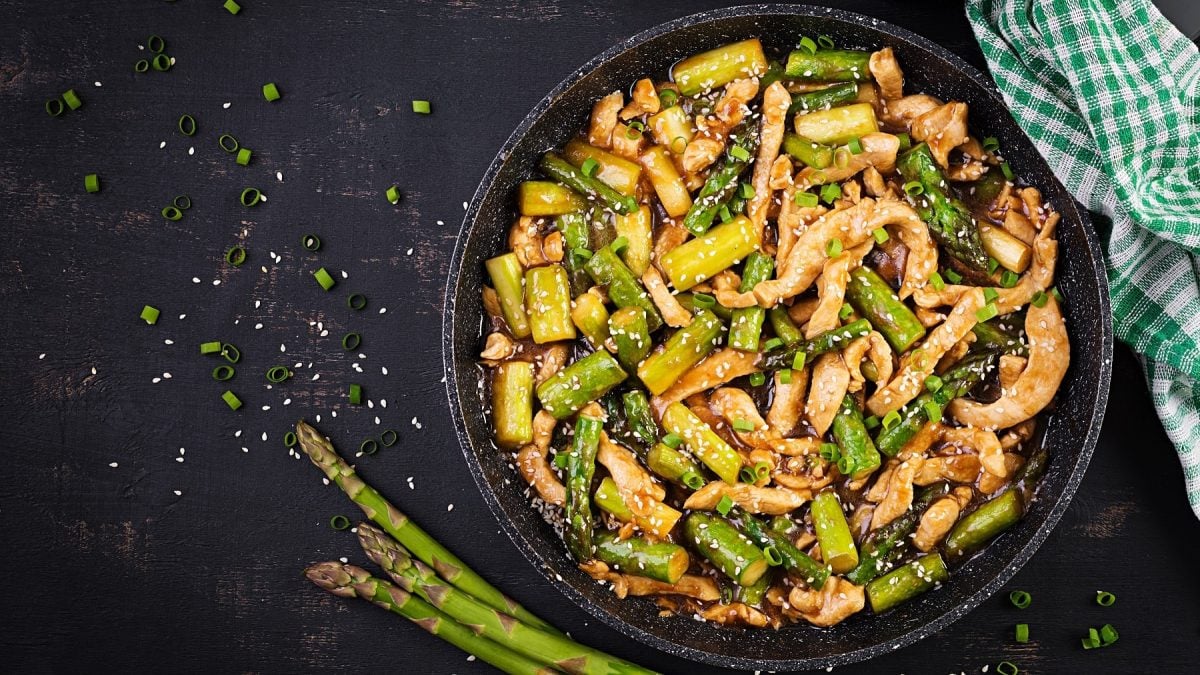 Eine schwarze Pfanne mit Spargel und Hähnchenstreifen auf einem schwarzen Untergrund. Daneben Sesam, Frühlingslauchringe und drei Stangen grüner Spargel. Direkt von oben fotografiert.