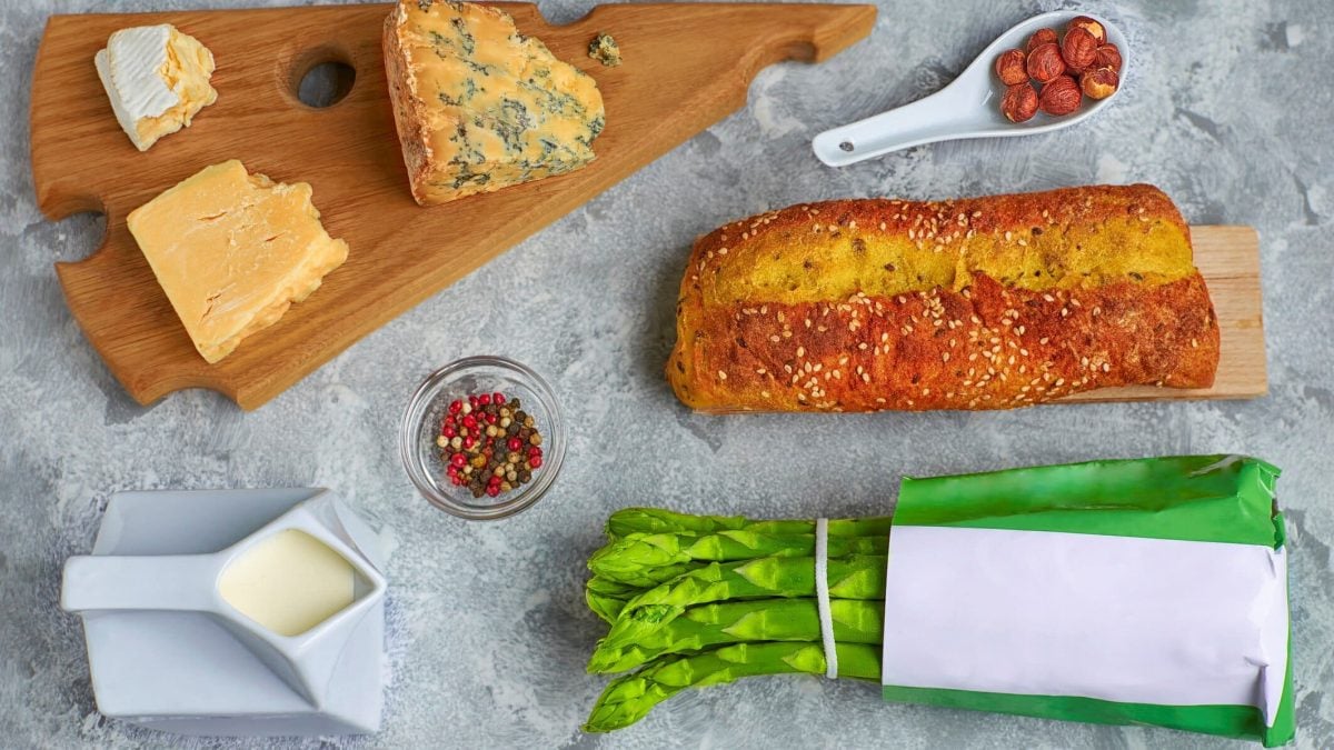 Spargelbot mit Grünem Spargel und Kurkuma auf Tisch mit Käseplatte, bunten Pfefferkörnern und einem Bund Grüner Spargel. Drausicht.