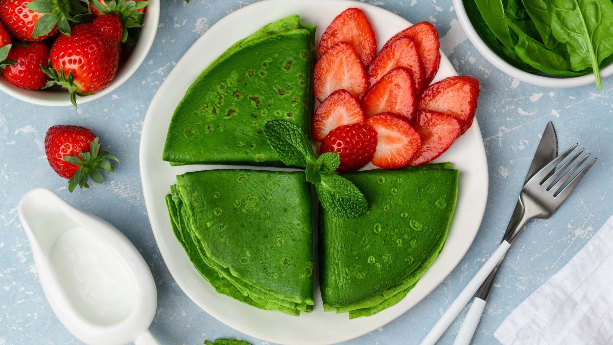 Draufsicht: auf einem blauen Untergrund steht ein Teller mit Spinat-Minze-Pfannkuchen mit Erdbeeren. Drumherum stehen eine Schale mit Erdbeeren, aufgeschnittenen Erdbeeren, ein Kännchen mit einer weißen Flüssigkeit sowie Messer und Gabel.