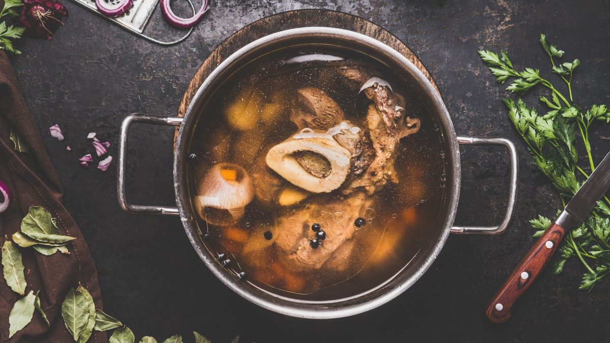 Draufsicht: Ein Topf selbst gemachte Rinderbrühe auf einen dunklen Holztisch. Drumherum liegen Kräuter, Lorbeerblätter, rote Zwiebelringe und ein Küchenmesser.