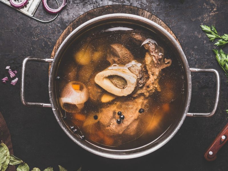 Draufsicht: Ein Topf selbst gemachte Rinderbrühe auf einen dunklen Holztisch. Drumherum liegen Kräuter, Lorbeerblätter, rote Zwiebelringe und ein Küchenmesser.