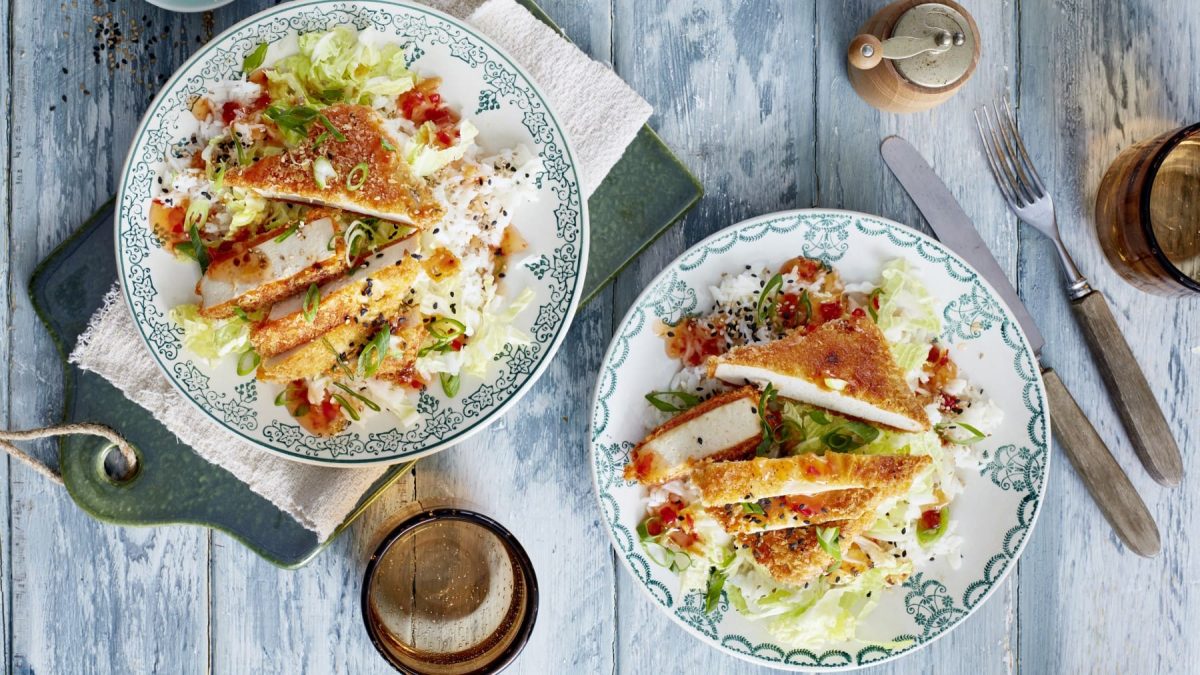 Zwei Teller mit angeschnittenen Tofu-Schnitzel mit Chinakohl neben Gewürzmühle, Glas und Besteck auf hellblauem Holzuntergrund.