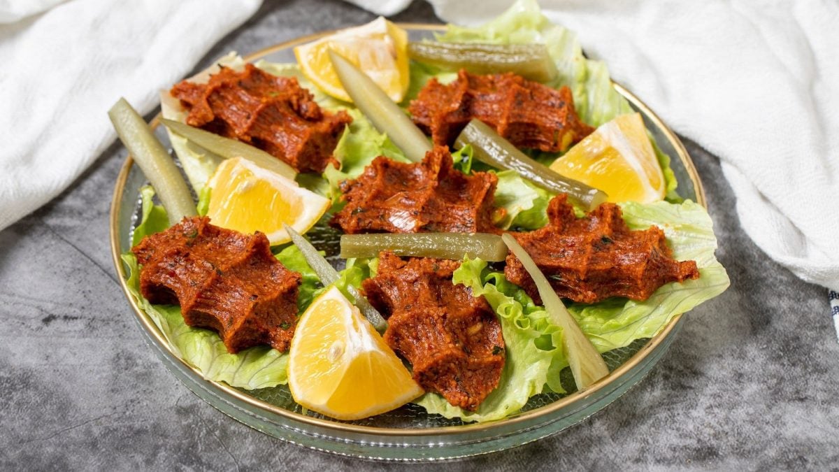 Vegane Çiğ Köfte auf Salatblättern mit Zitronenstücken und in Streifen geschnittener Gewürzgurken auf einem silbernen Teller.